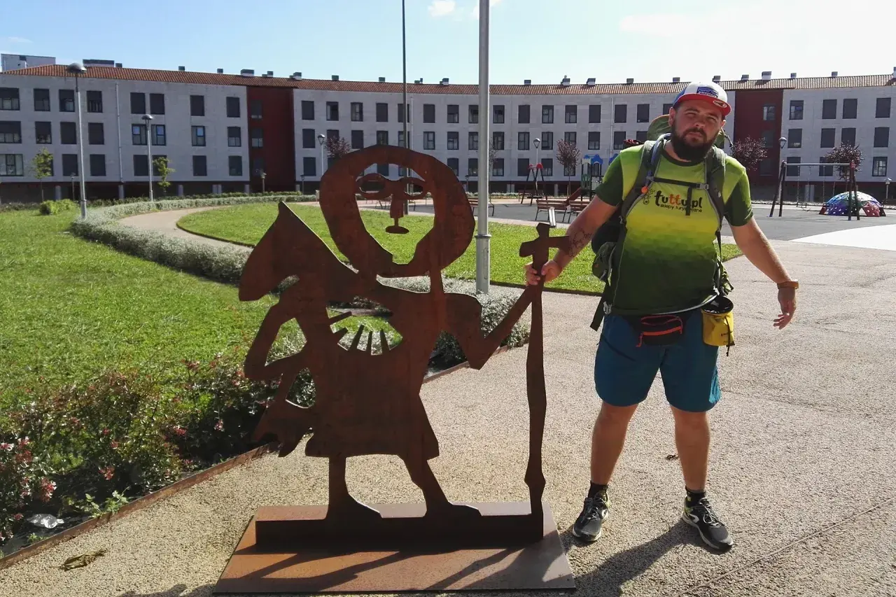 Pielgrzym na camino de santiago pozuje obok rdzewiejącej rzeźby pielgrzyma.
