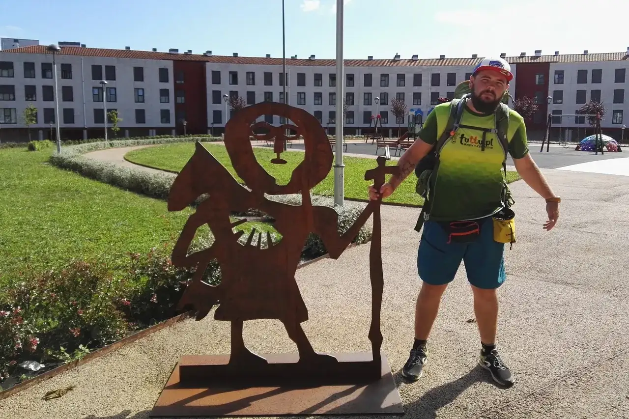 Pielgrzym na camino de santiago pozuje obok rdzewiejącej rzeźby pielgrzyma.